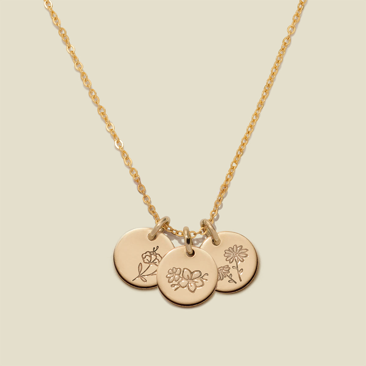 A gold chain necklace with three round engraved gold disc pendants featuring delicate floral designs, displayed against a solid light beige background.