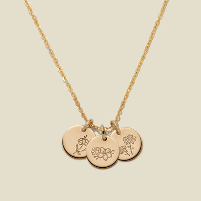 A gold chain necklace with three round engraved gold disc pendants featuring delicate floral designs, displayed against a solid light beige background.