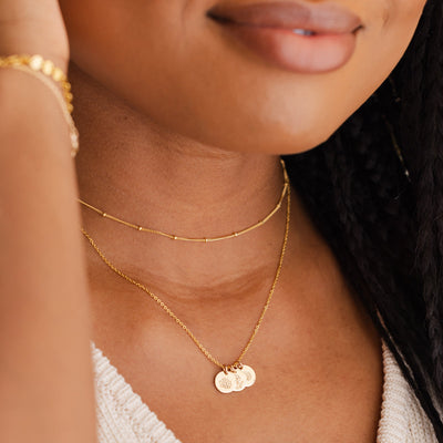 A close-up of a woman wearing a delicate gold chain necklace with tiny round stationary beads, layered with a gold pendant chain featuring three round flat discs engraved with various floral designs.