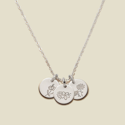 A silver chain necklace with three round engraved silver disc pendants with floral designs, on a light beige background.