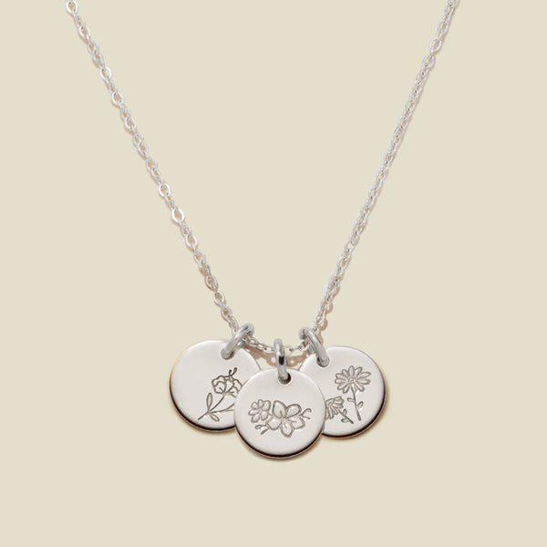 A silver chain necklace with three round engraved silver disc pendants with floral designs, on a light beige background.