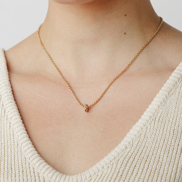 A close-up lifestyle image of a person wearing a gold chain chain necklace with a small ring charm in an eternity style with blue square gemstones, set against a white background.