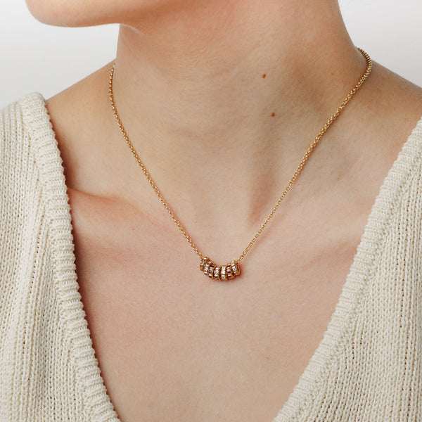 A close-up lifestyle image of a person wearing a gold chain chain necklace with seven small ring charms, each with a different color of gemstone, set against a white background.