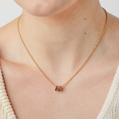A close-up lifestyle image of a person wearing a gold chain chain necklace with three small ring charms, each with a different color of gemstone, set against a white background.