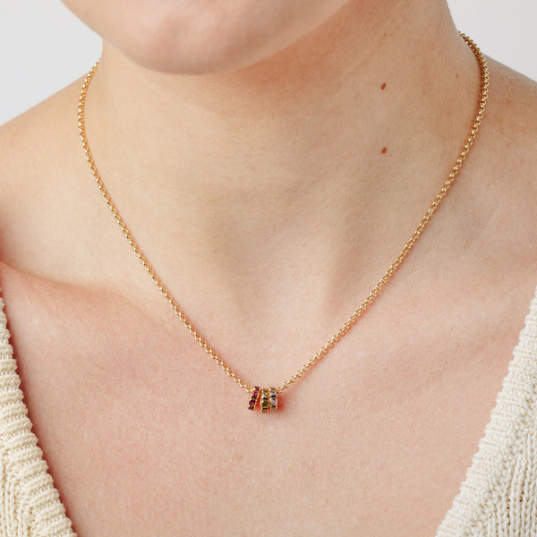 A close-up lifestyle image of a person wearing a gold chain chain necklace with three small ring charms, each with a different color of gemstone, set against a white background.