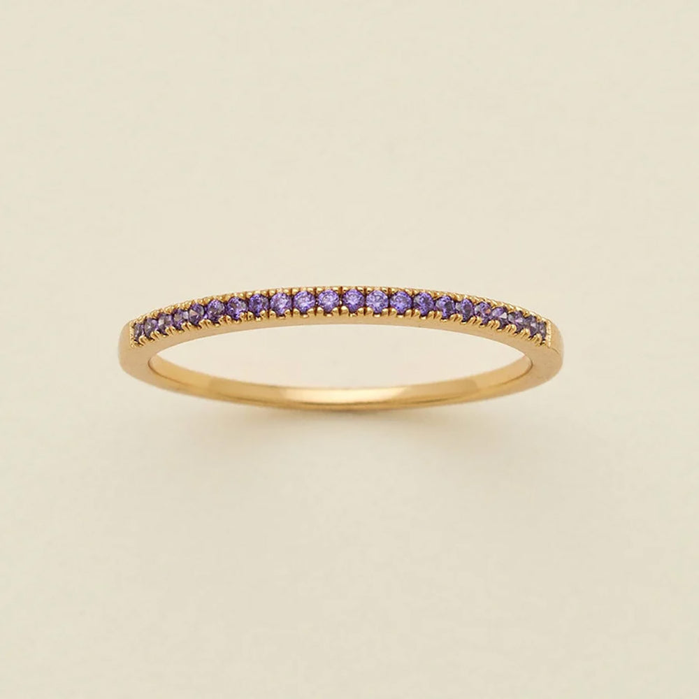 A gold ring featuring a single row of small round-cut dark purple gemstones, photographed on a clean, solid, warm off-white background.