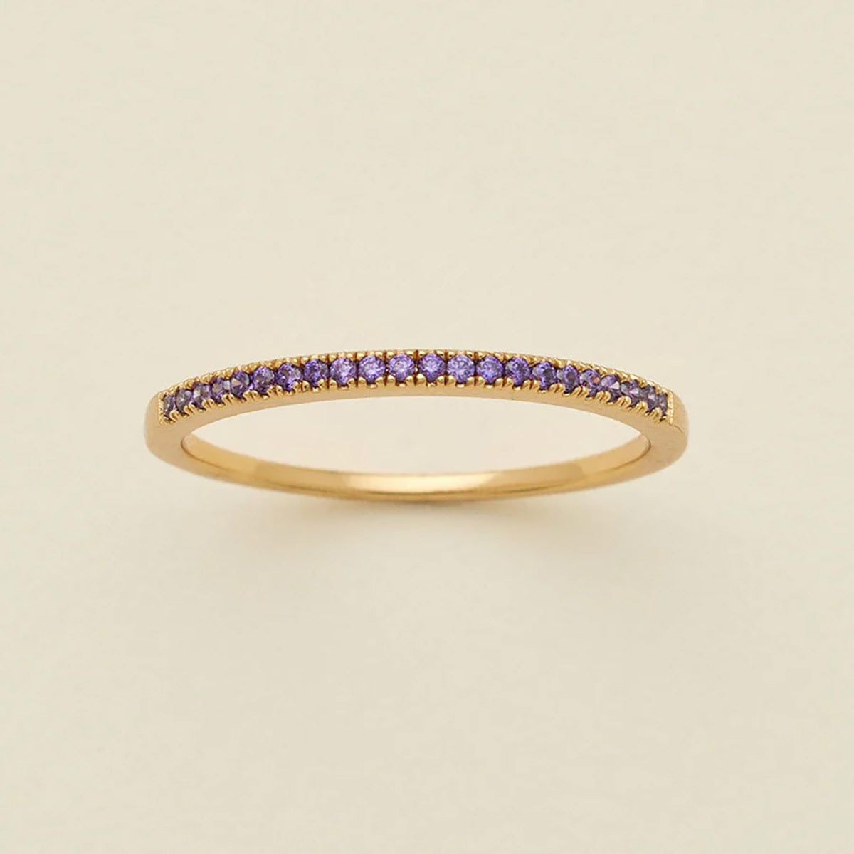 A gold ring featuring a single row of small round-cut dark purple gemstones, photographed on a clean, solid, warm off-white background.