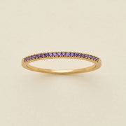 A gold ring featuring a single row of small round-cut dark purple gemstones, photographed on a clean, solid, warm off-white background.