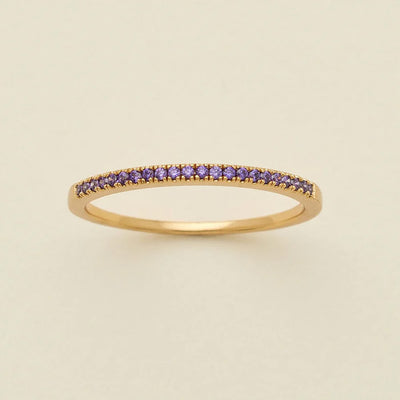 A gold ring featuring a single row of small round-cut dark purple gemstones, photographed on a clean, solid, warm off-white background.