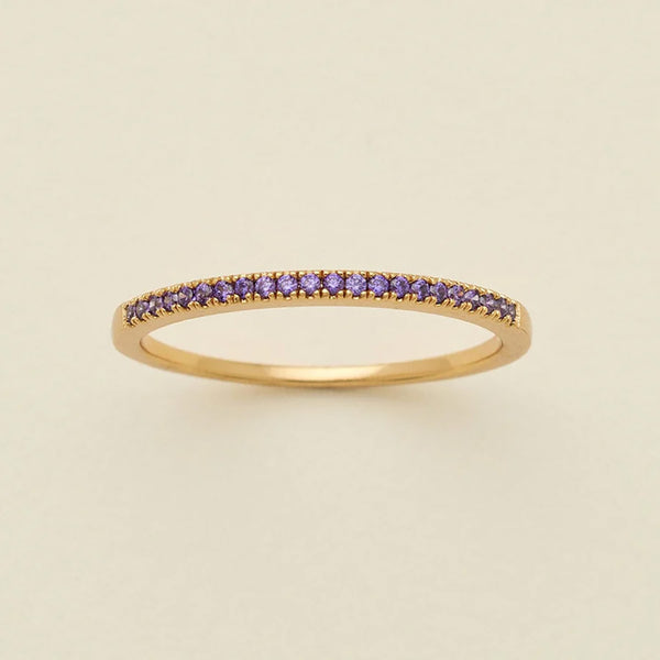 A gold ring featuring a single row of small round-cut dark purple gemstones, photographed on a clean, solid, warm off-white background.