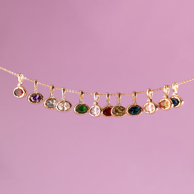 A dainty gold chain pulled horizontally featuring multiple gold oval gemstone charms in varying colors, set against a pink background.