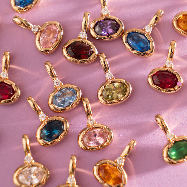 Multiple gold oval pendant charms with differing oval gemstone colors, laid on a pink background.