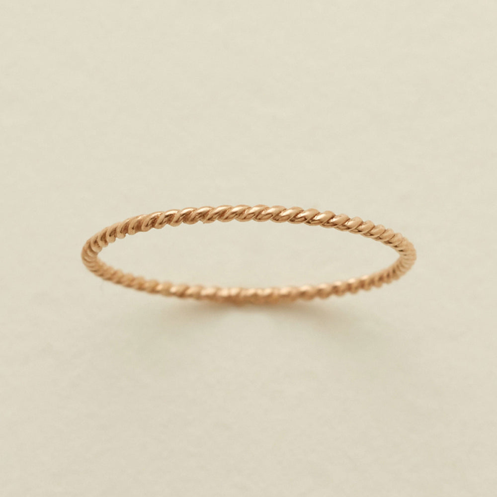 A thin, twisted rope-style ring in a warm gold metal, photographed on a clean, light beige textured background.