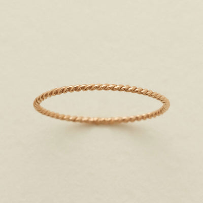 A thin, twisted rope-style ring in a warm gold metal, photographed on a clean, light beige textured background.