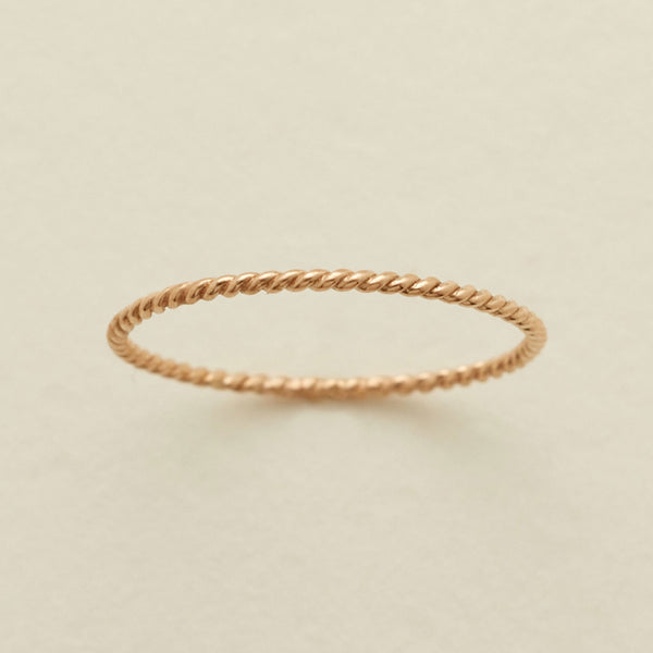 A thin, twisted rope-style ring in a warm gold metal, photographed on a clean, light beige textured background.