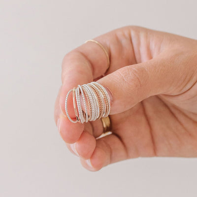 A hand holding multiple gold and silver thin, twisted rope wire rings on their thumb, shown on a solid white background.