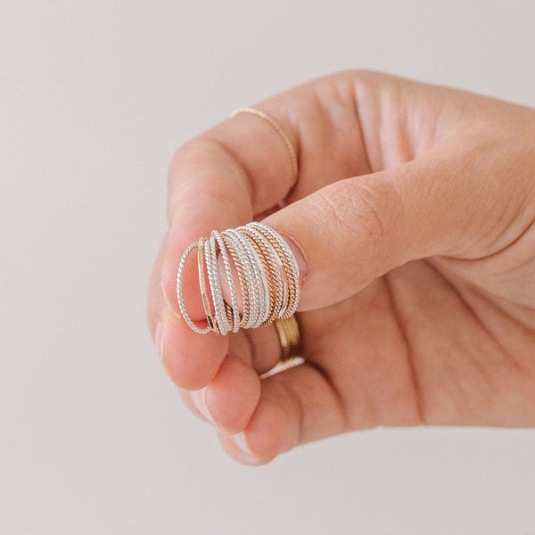 A hand holding multiple gold and silver thin, twisted rope wire rings on their thumb, shown on a solid white background.