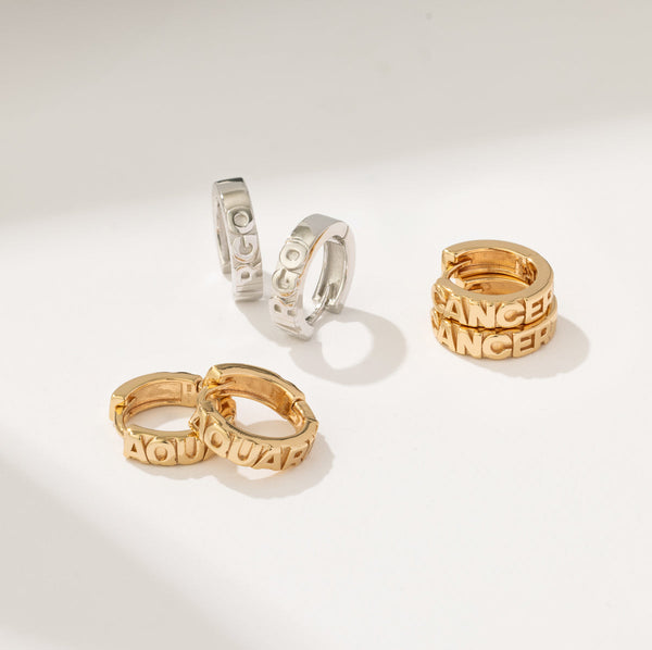 Three pairs of huggie hoop earrings, two gold and one silver, each with a different zodiac sign in raised letters across the front and set on a solid white surface.