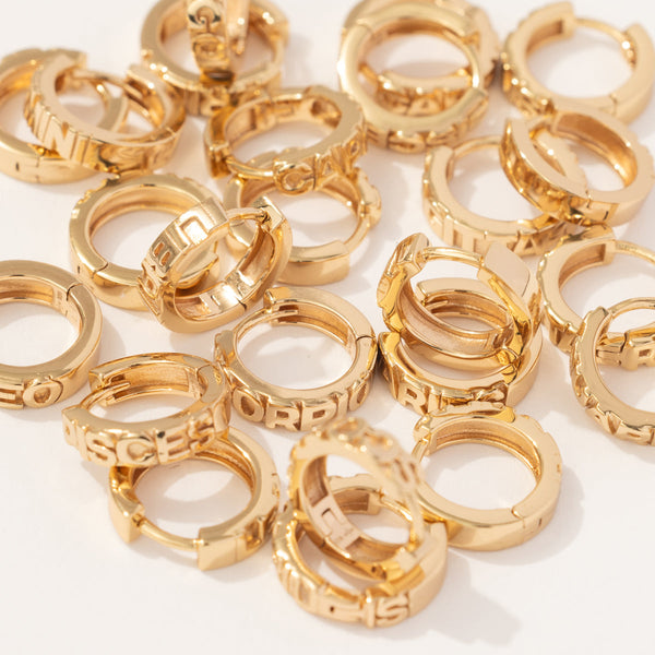 Multiple gold huggie hoop earrings scattered on a solid white surface. Each hoop has a different name of a zodiac sign in raised letters across the front.