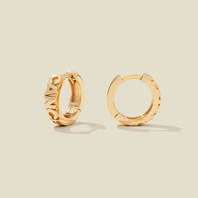 A pair of gold huggie hoop earrings on a light beige background. One earring is shown from the side, displaying a polished outer surface, while the other is shown from the front, showcasing the word 'CANCER' in raised letters.