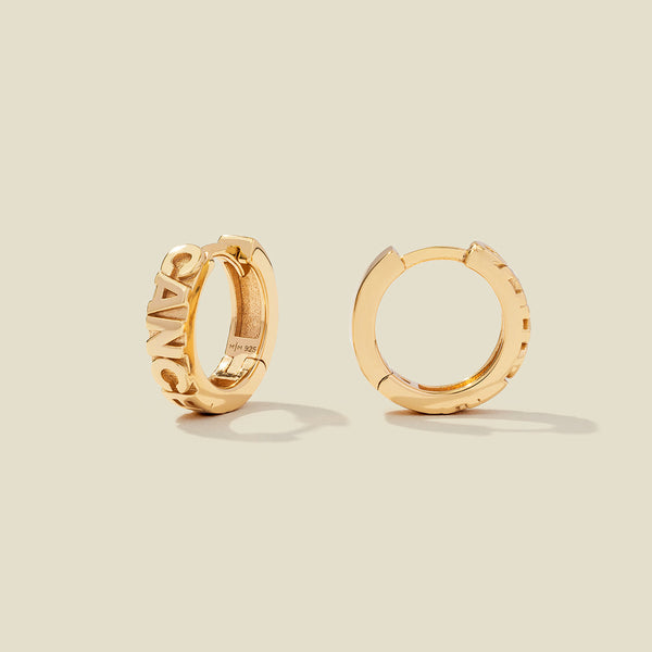 A pair of gold huggie hoop earrings on a light beige background. One earring is shown from the side, displaying a polished outer surface, while the other is shown from the front, showcasing the word 'CANCER' in raised letters.