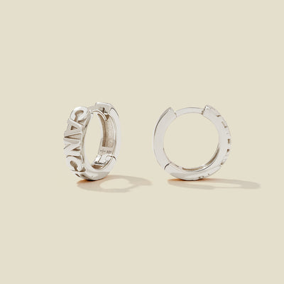 A pair of silver huggie hoop earrings on a light beige background. One earring is shown from the side, displaying a polished outer surface, while the other is shown from the front, showcasing the word 'CANCER' in raised letters.