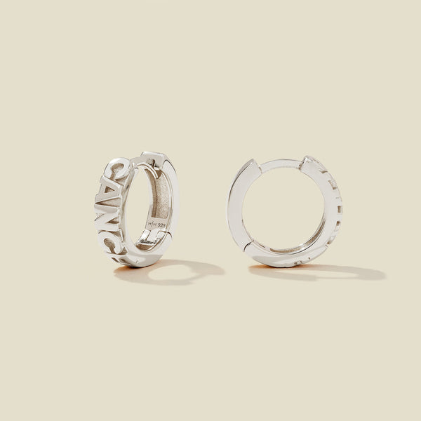 A pair of silver huggie hoop earrings on a light beige background. One earring is shown from the side, displaying a polished outer surface, while the other is shown from the front, showcasing the word 'CANCER' in raised letters.