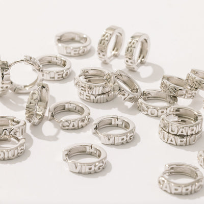 Multiple silver huggie hoop earrings laying scattered on a solid white background. Each of the hoops features the name of a zodiac sign along the front in raised letters.
