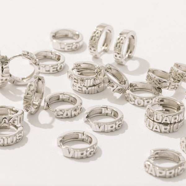 Multiple silver huggie hoop earrings laying scattered on a solid white background. Each of the hoops features the name of a zodiac sign along the front in raised letters.