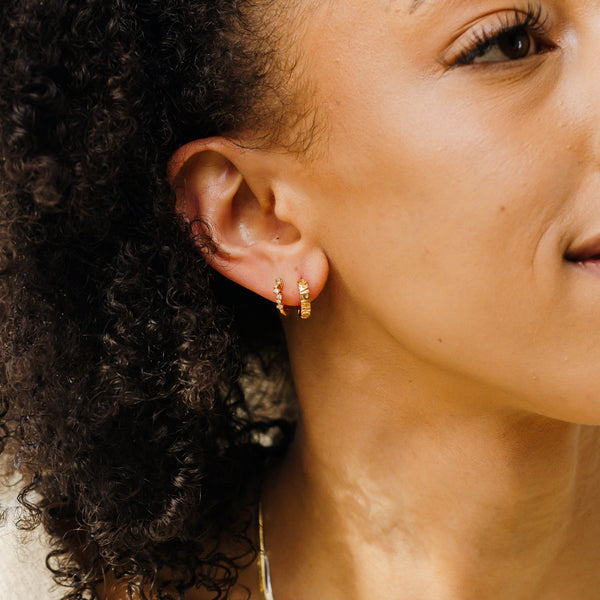 A close-up of a woman's ear wearing two gold hoop earrings, one with round clear gemstones in a curving pattern and the other with the word 'CAPRICORN' down the front in raised letters.