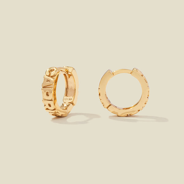 A pair of gold huggie hoop earrings on a light beige background. One earring is shown from the side, displaying a polished outer surface, while the other is shown from the front, showcasing the word 'CAPRICORN' in raised letters.