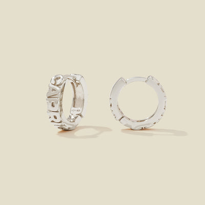A pair of silver huggie hoop earrings on a light beige background. One earring is shown from the side, displaying a polished outer surface, while the other is shown from the front, showcasing the word 'CAPRICORN' in raised letters.