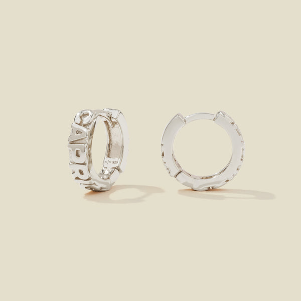 A pair of silver huggie hoop earrings on a light beige background. One earring is shown from the side, displaying a polished outer surface, while the other is shown from the front, showcasing the word 'CAPRICORN' in raised letters.