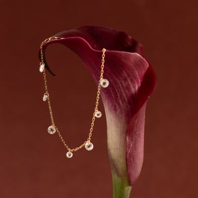 A gold chain bracelet with dangling clear crystal stones, set on a red flower against a warm maroon backdrop.