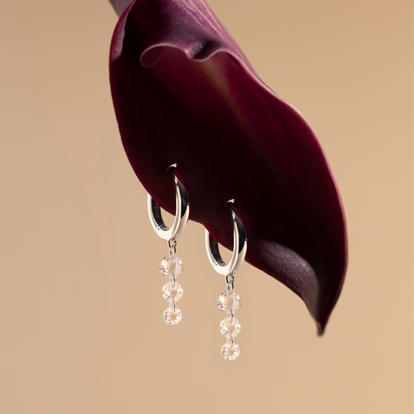 Two silver hoop earrings with clear gemstones hanging vertically from a purple flower petal on a beige background