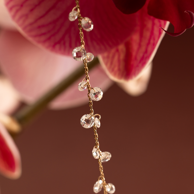 A dainty gold necklace with clusters of dangling clear crystals hanging from a dark pink flower against a maroon background.