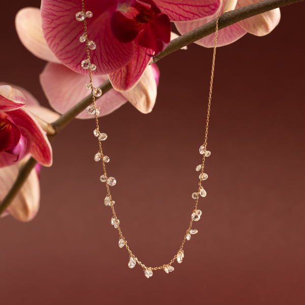 A dainty gold necklace with clusters of dangling clear crystals hanging from a dark pink flower against a maroon background.