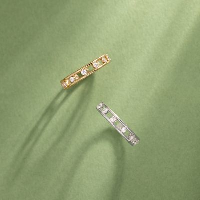 A gold and a silver eternity ring with marquise-cut clear stones set between two thin bands, with open space between, set against a light green background with natural shadows.