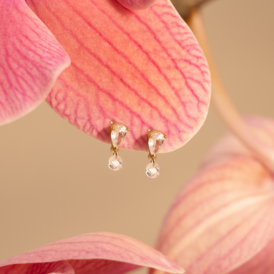 A pair of gold stud earrings with upside down tear-drop shaped clear stones, and a small clear crystal dangling from the point, set on a pink flower petal against a beige backdrop.