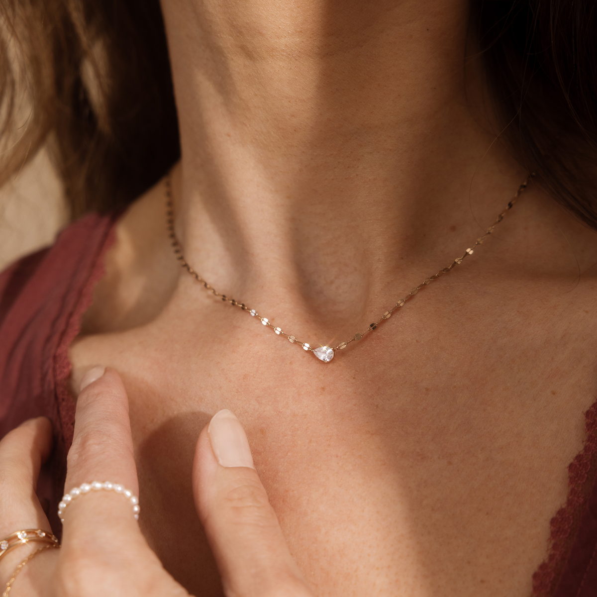 A close up lifestyle shot of a person's neck wearing a dainty gold chain necklace featuring a sideways tear-drop shaped clear stone in the center. The person is also wearing a pearl eternity ring, styled with a red knit blouse.