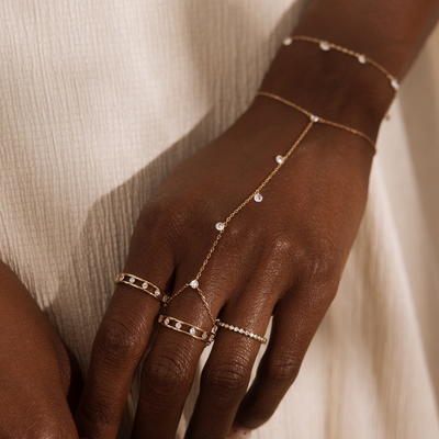 A close up lifestyle shot of an arm and hand with multiple gold rings with clear stones in different styles, a gold hand chain with dangling clear stones, and a matching gold bracelet.