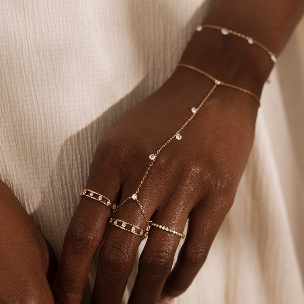 A close up lifestyle shot of an arm and hand with multiple gold rings with clear stones in different styles, a gold hand chain with dangling clear stones, and a matching gold bracelet.