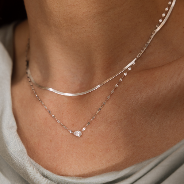 A close up lifestyle shot of two layered silver necklaces on a person's neck, one minimalistic shorter style and a longer textured chain featuring a sideways tear-drop shaped clear stone.