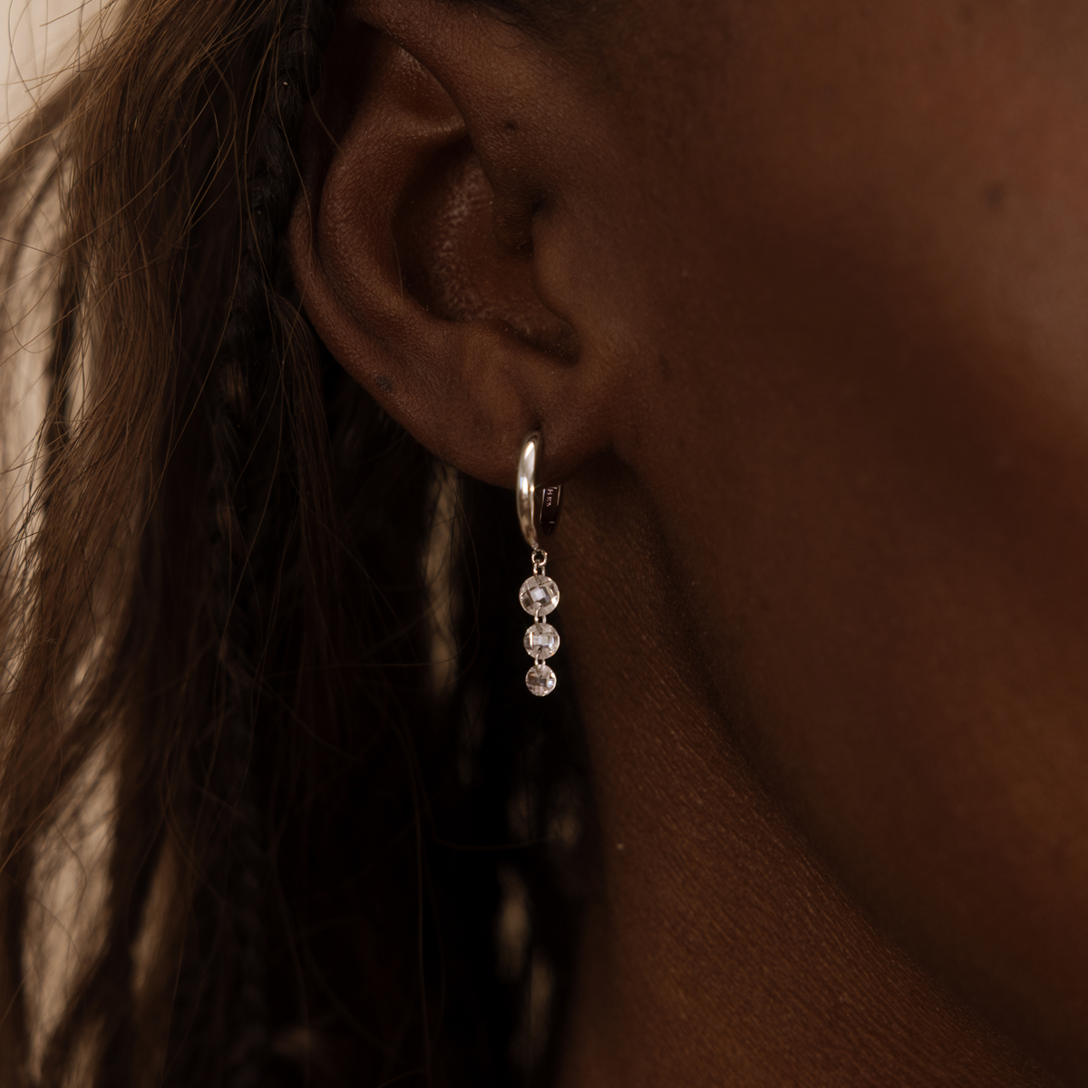 Close-up of an ear wearing a silver hoop earring with three clear stones hanging vertically.