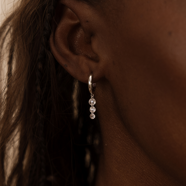 Close-up of an ear wearing a silver hoop earring with three clear stones hanging vertically.