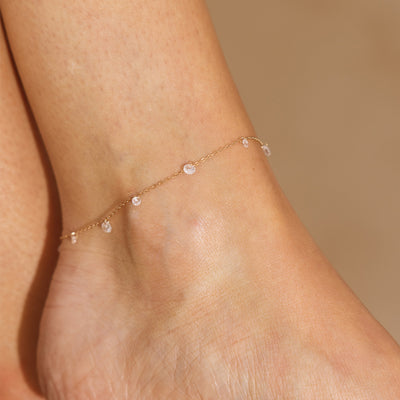 Gold anklet with small clear dangling stones on a person's ankle against a beige background