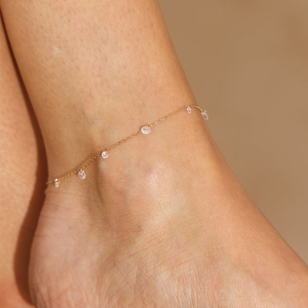 Gold anklet with small clear dangling stones on a person's ankle against a beige background