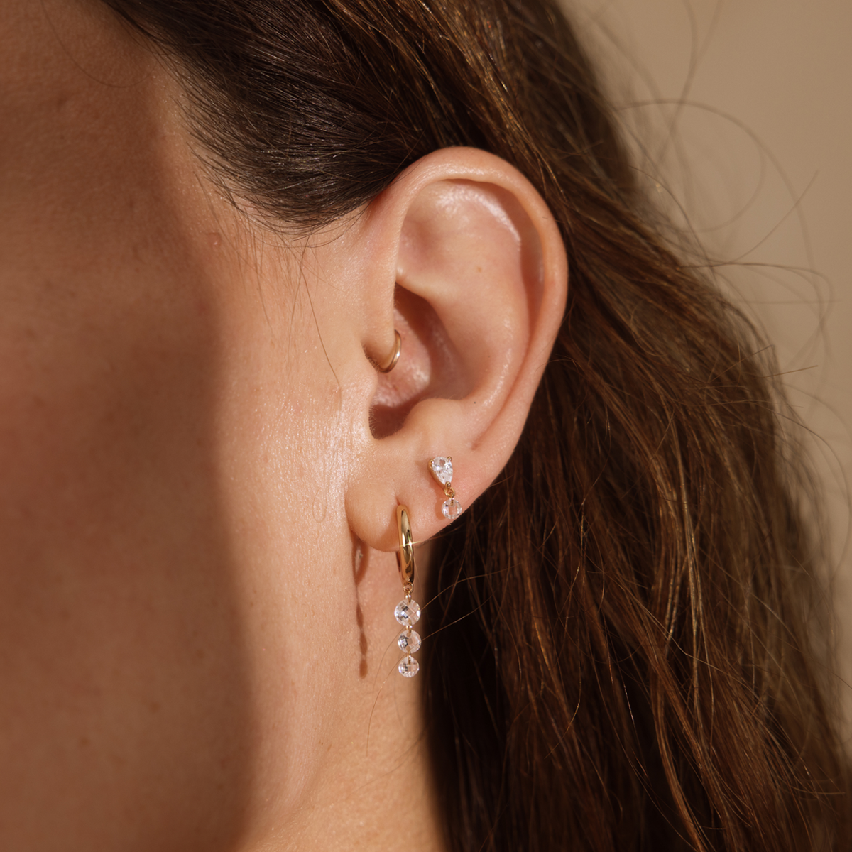 A close up lifestyle shot of an ear wearing a gold hoop with dangling clear stones, a stud with two clear stones, and a thin gold hoop, against a beige background.