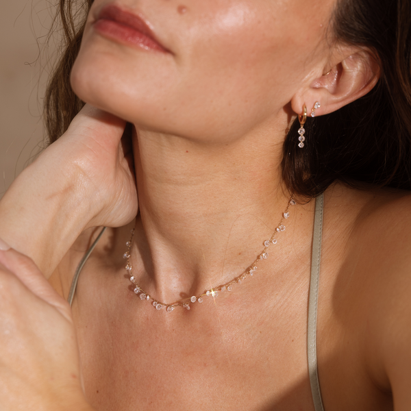 A close up lifestyle shot of a person's neck wearing a gold chain necklace with clusters of round clear crystals dangling down, styled with a gold hoop with three hanging round clear crystals.
