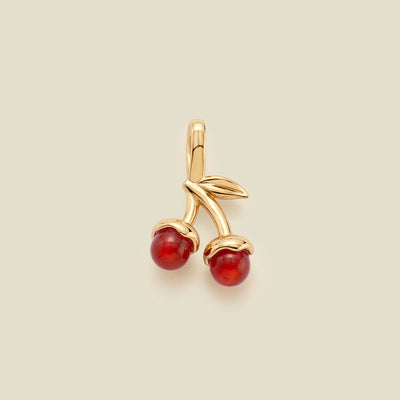 A cherry-shaped jewelry charm in polished gold-tone metal with two red agate stone cherries and small gold leaves, centered on a light beige background.